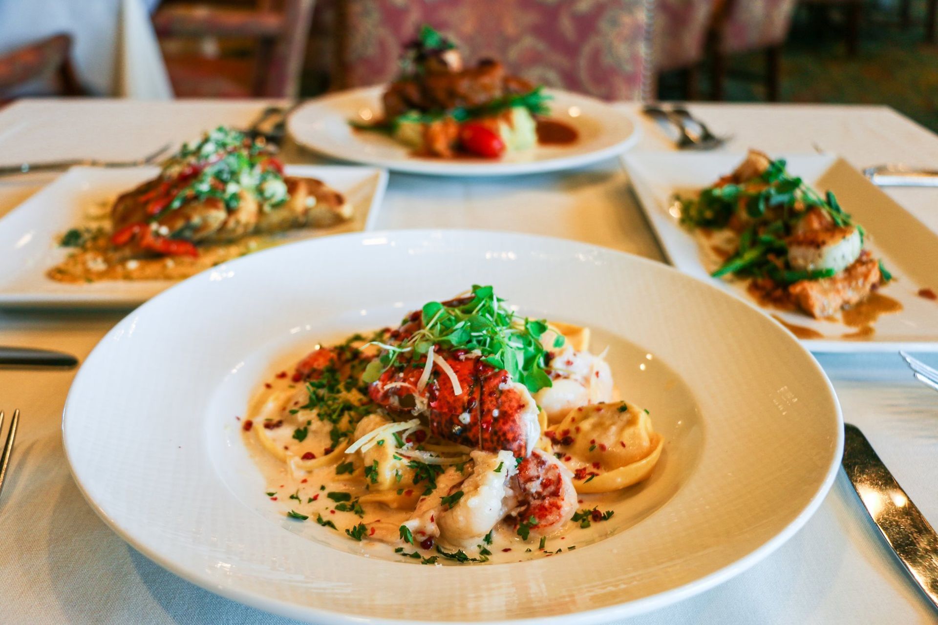 Four gourmet dishes on a white tablecloth; lobster pasta in foreground, other entrees behind.