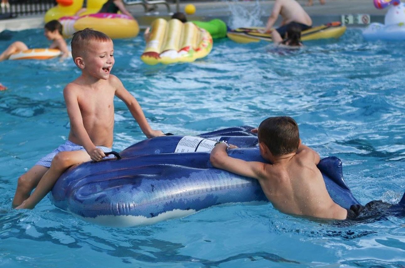 Two boys laugh while playing on a blue inflatable whale in a pool with other people.