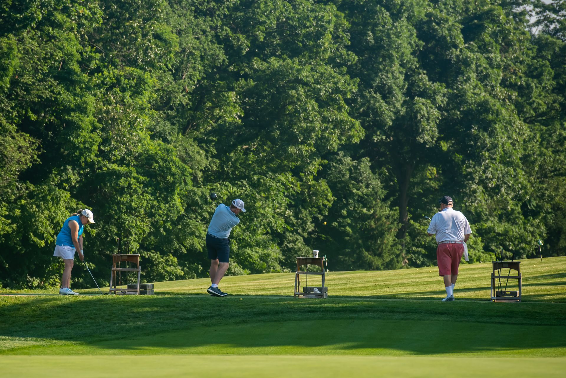 Three golfers teeing off on a green fairway beside a tree-lined course