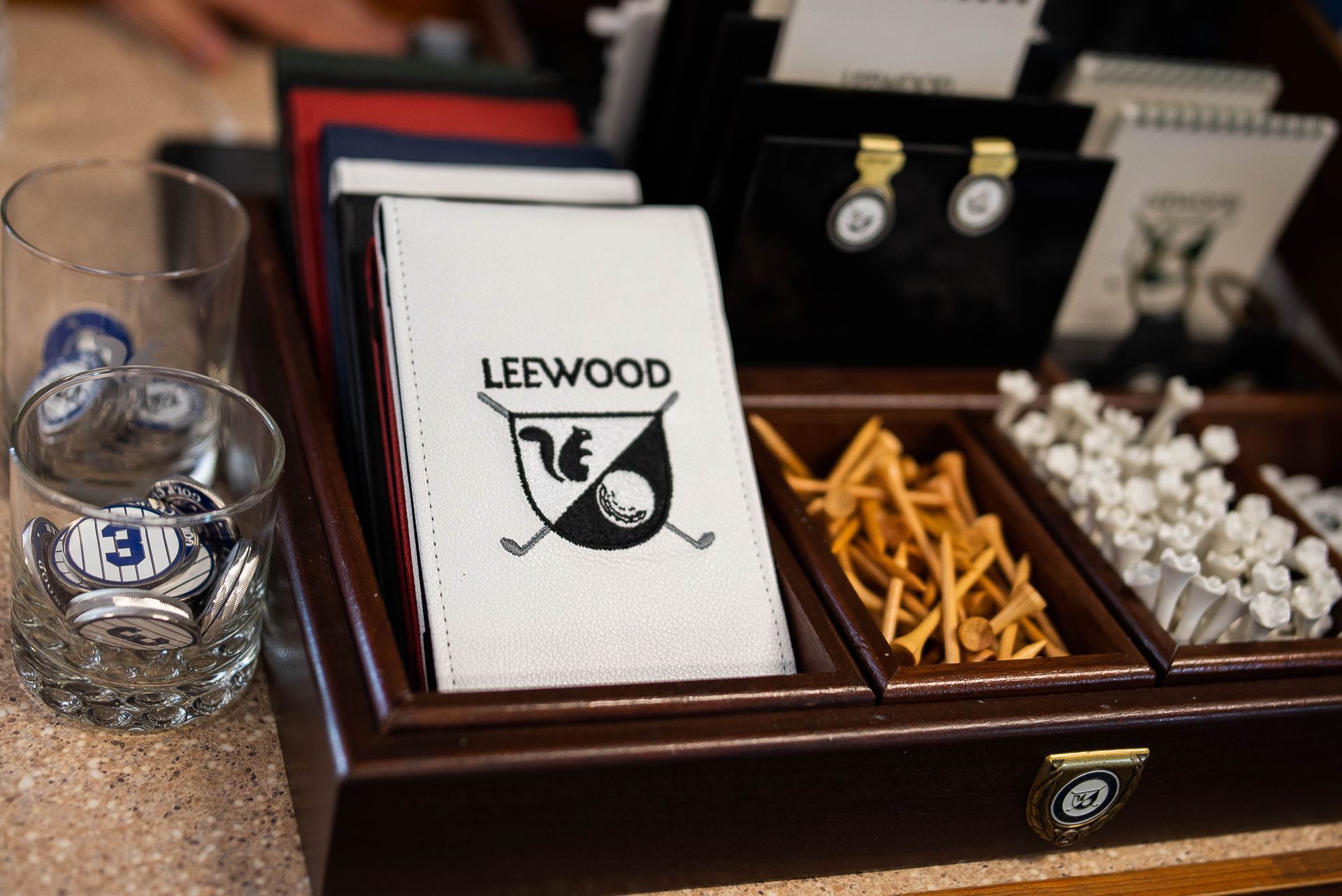 A wooden organizer containing a Leewood golf scorecard cover, wooden golf tees, white plastic tees, and branded glasses.