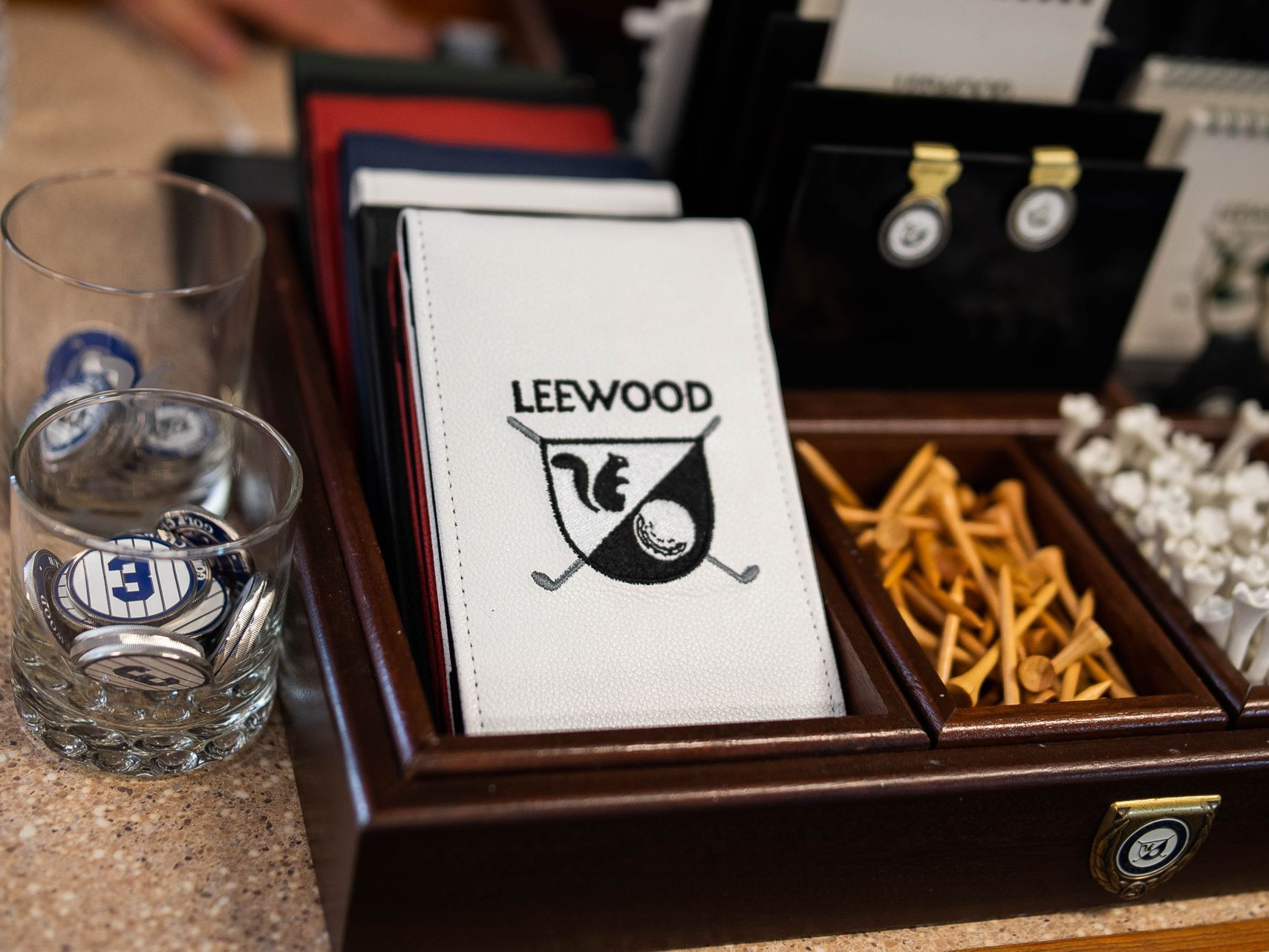 A wooden organizer containing a Leewood golf scorecard cover, wooden golf tees, white plastic tees, and branded glasses.