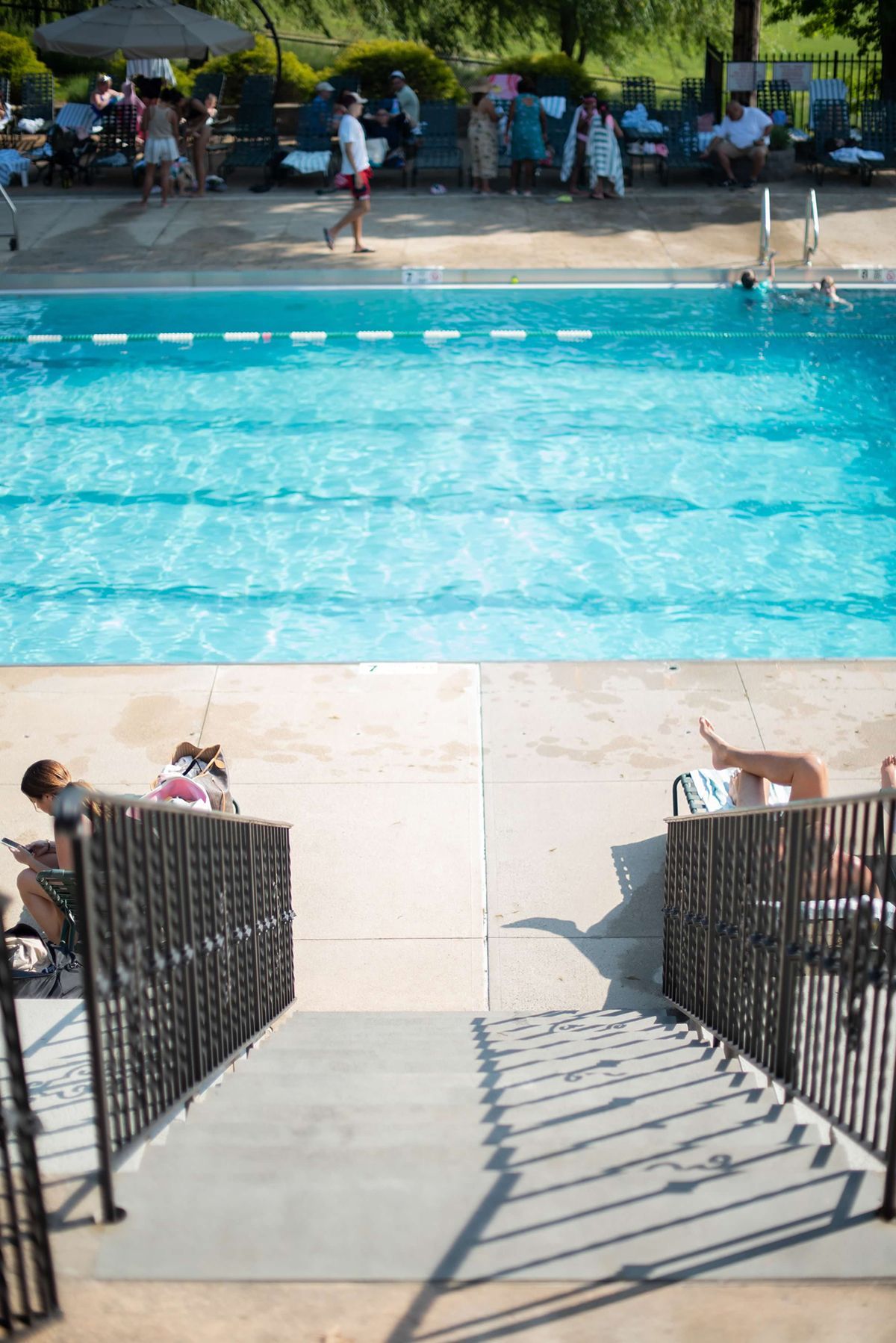 Pool with swimmers, lounge chairs, and people in the background. Stairs with railing in foreground.
