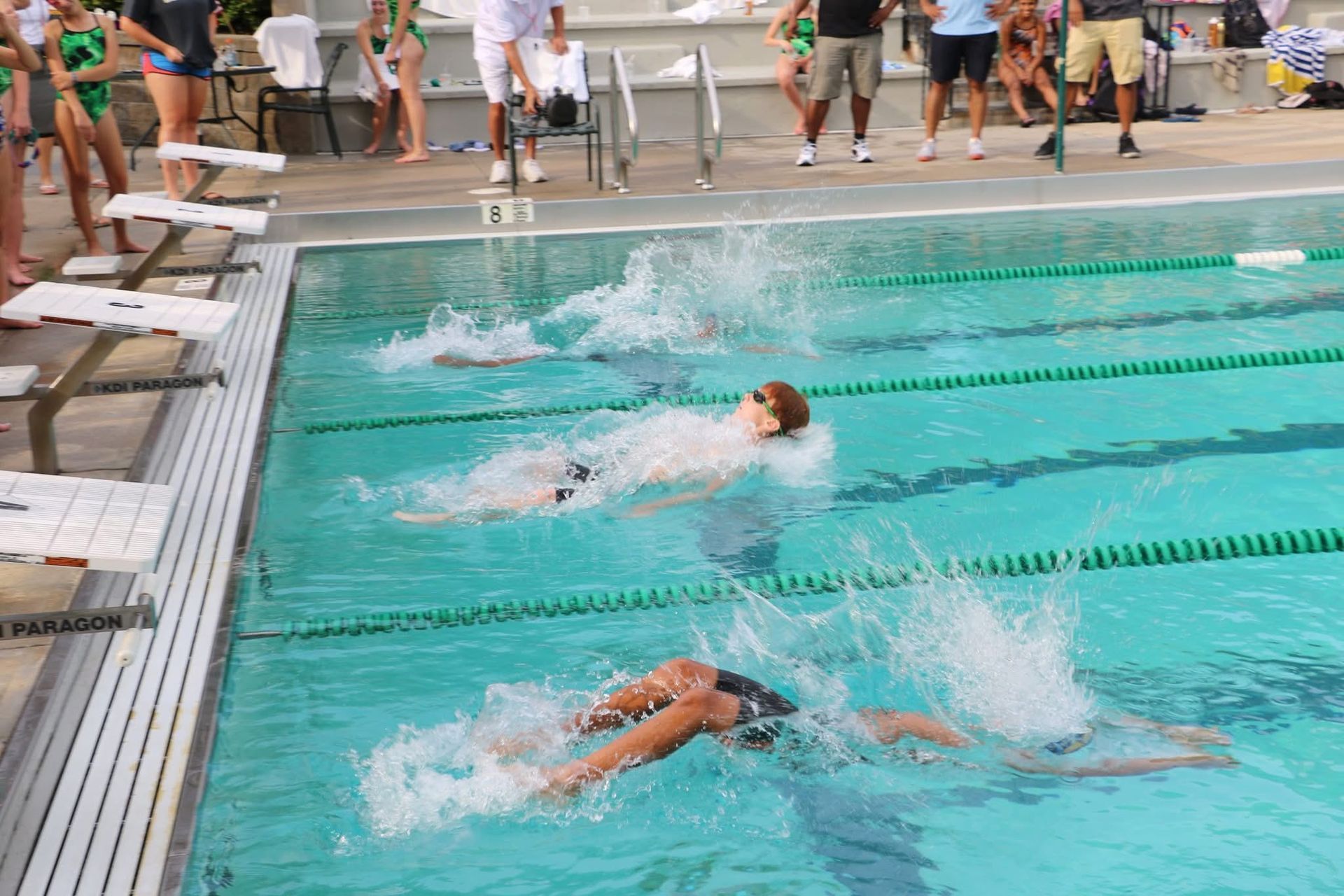 Swimmers in a pool, mid-stroke, water splashing. People watch from the pool's edge.