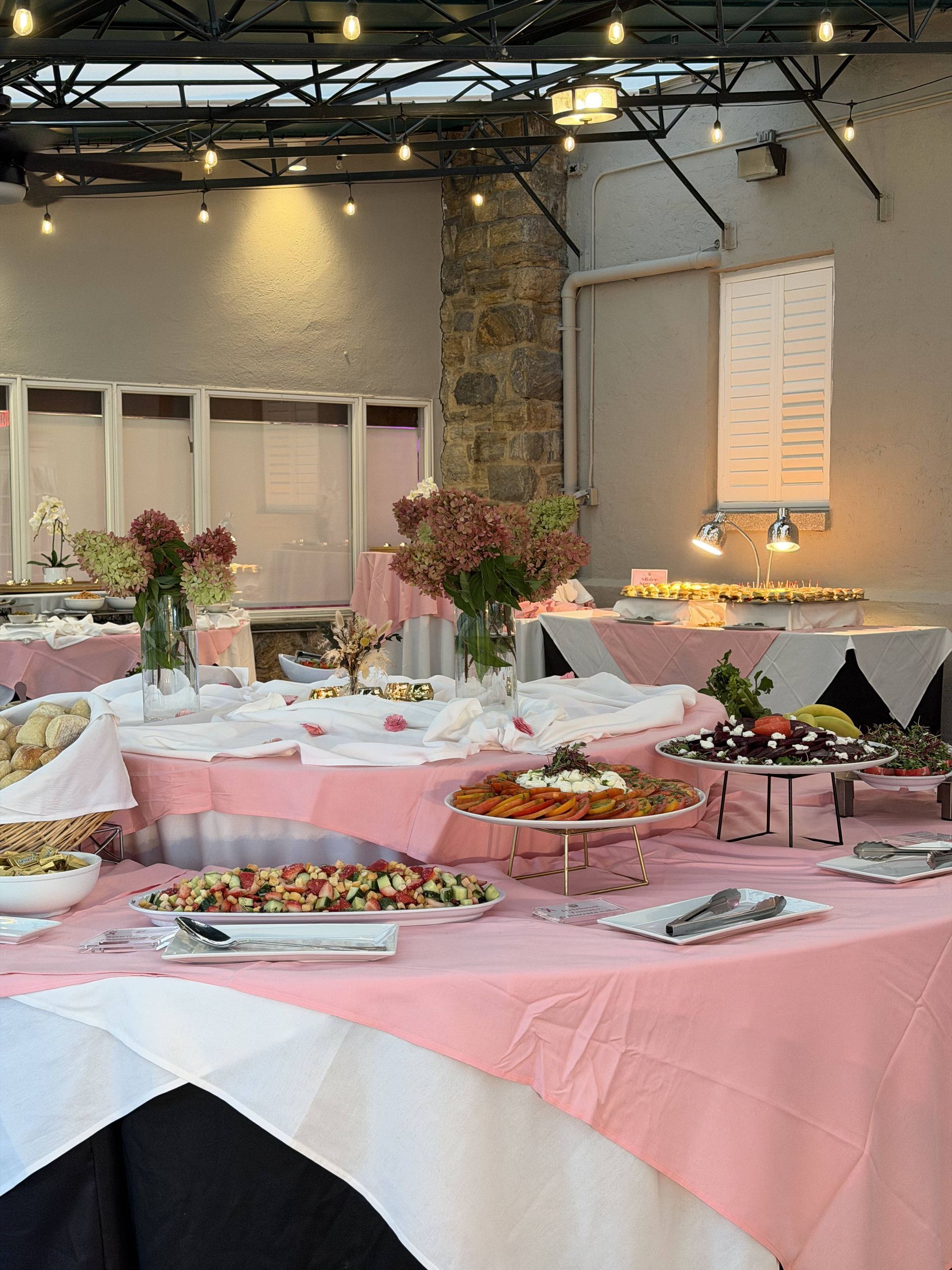 Buffet table with pink tablecloths, floral arrangements, and trays of food in a decorated indoor venue