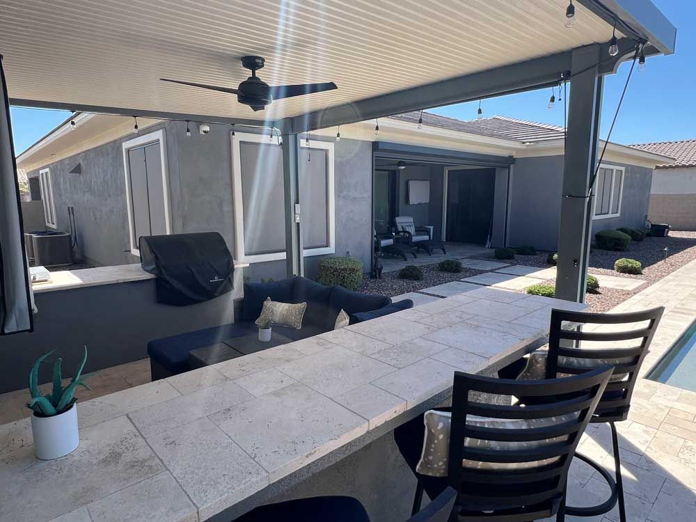 A patio area with a bar, chairs, a grill and a ceiling fan.