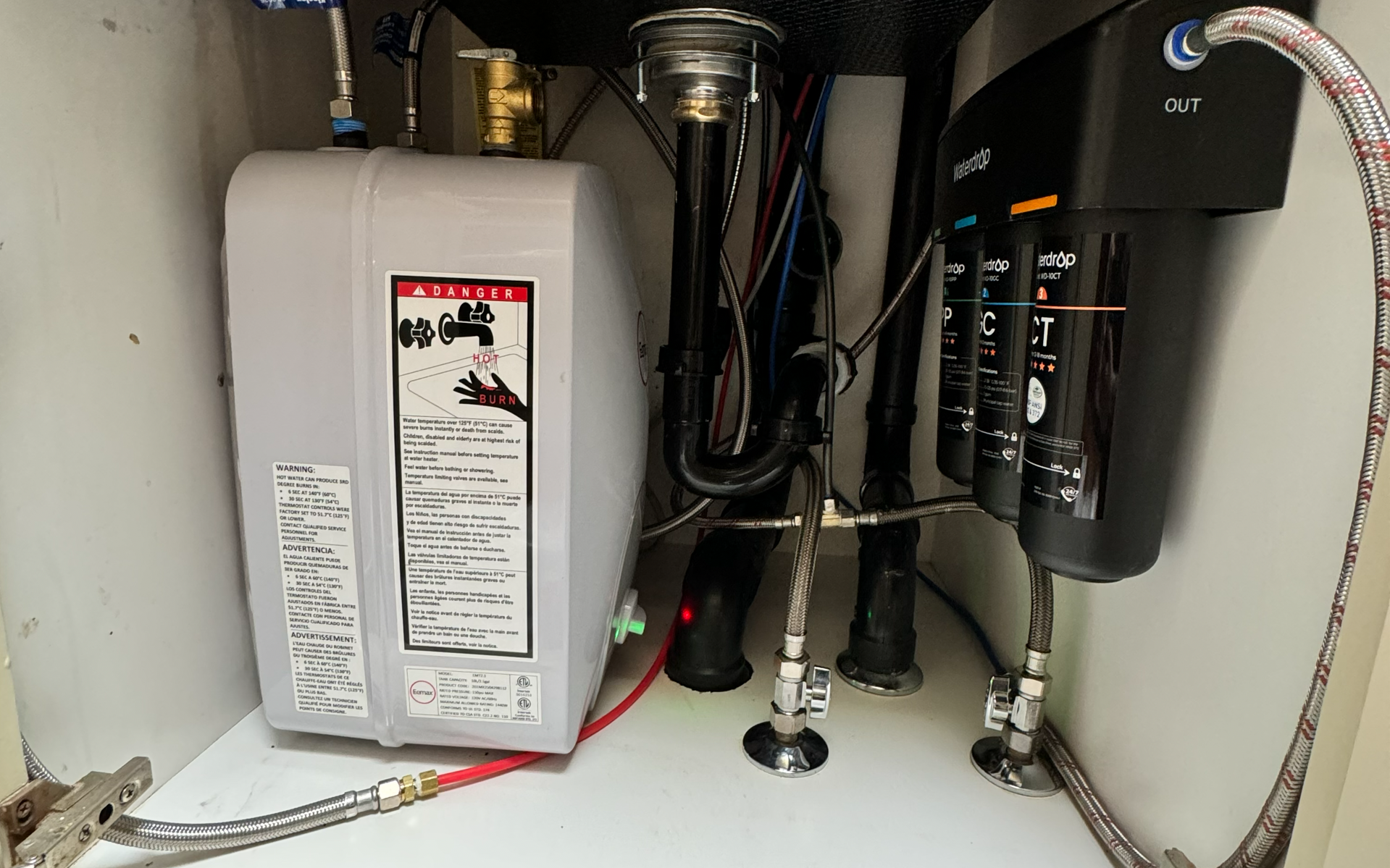 Under-sink plumbing with water heater, pipes, and black filtration unit in a cabinet
