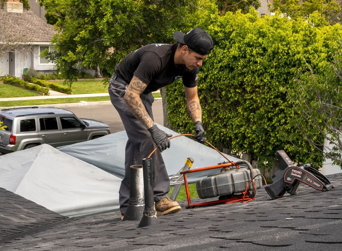 Worker on a roof installing shingles beside tools and a rolled tarp.