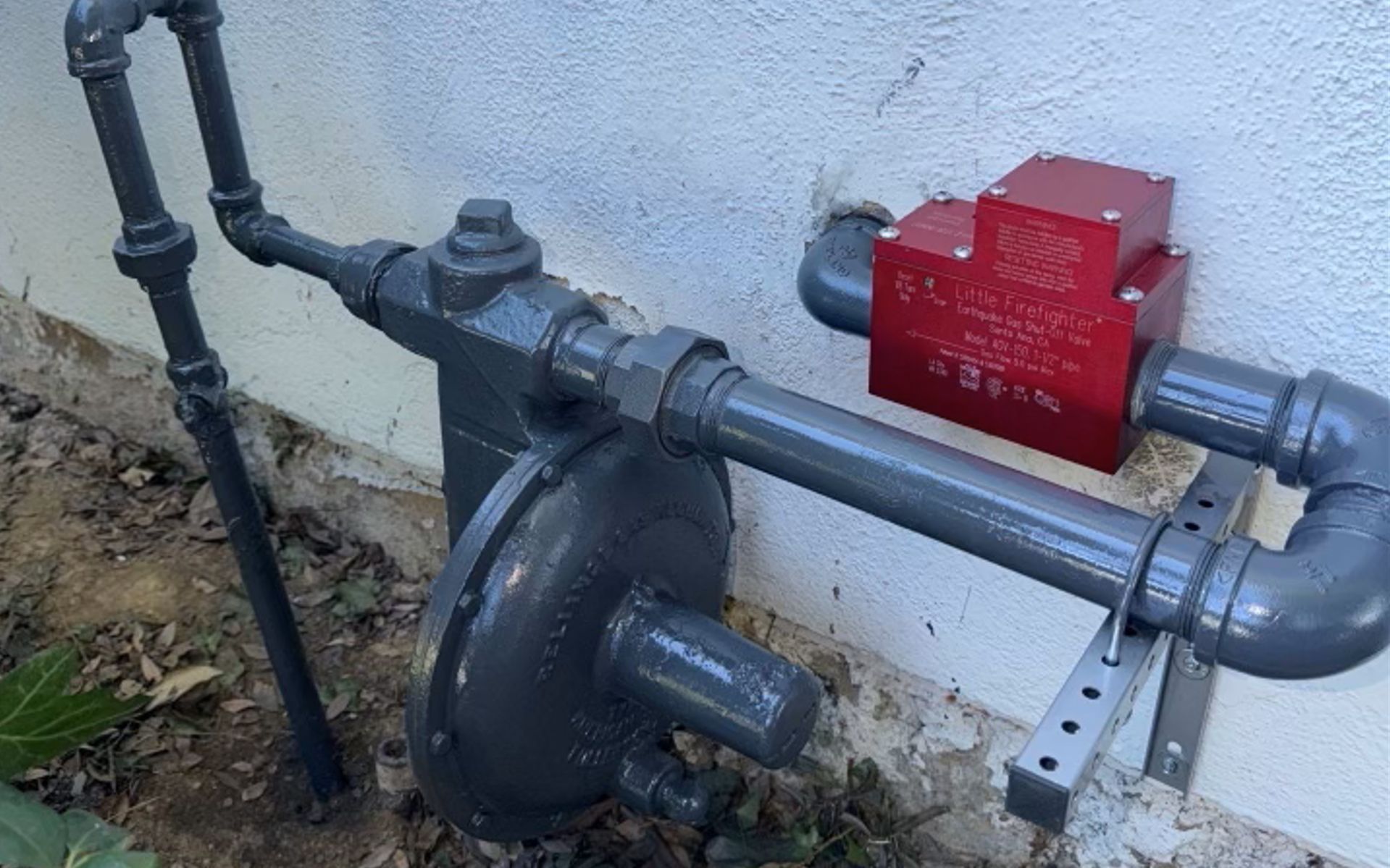 Outdoor utility pipes with a red gas meter/regulator mounted on a wall.