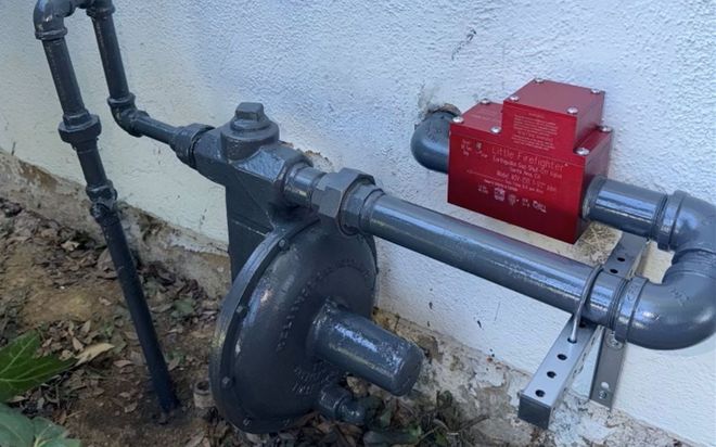 Outdoor utility pipes with a red gas meter/regulator mounted on a wall.