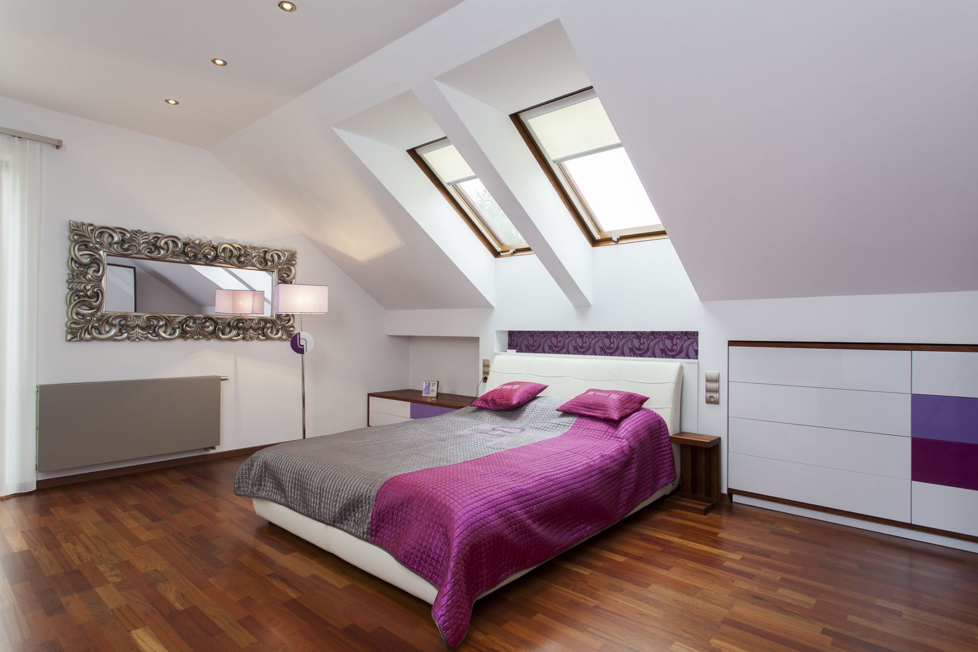 Bedroom with slanted ceiling, skylights, white walls, wood floor, bed with purple and grey bedding.