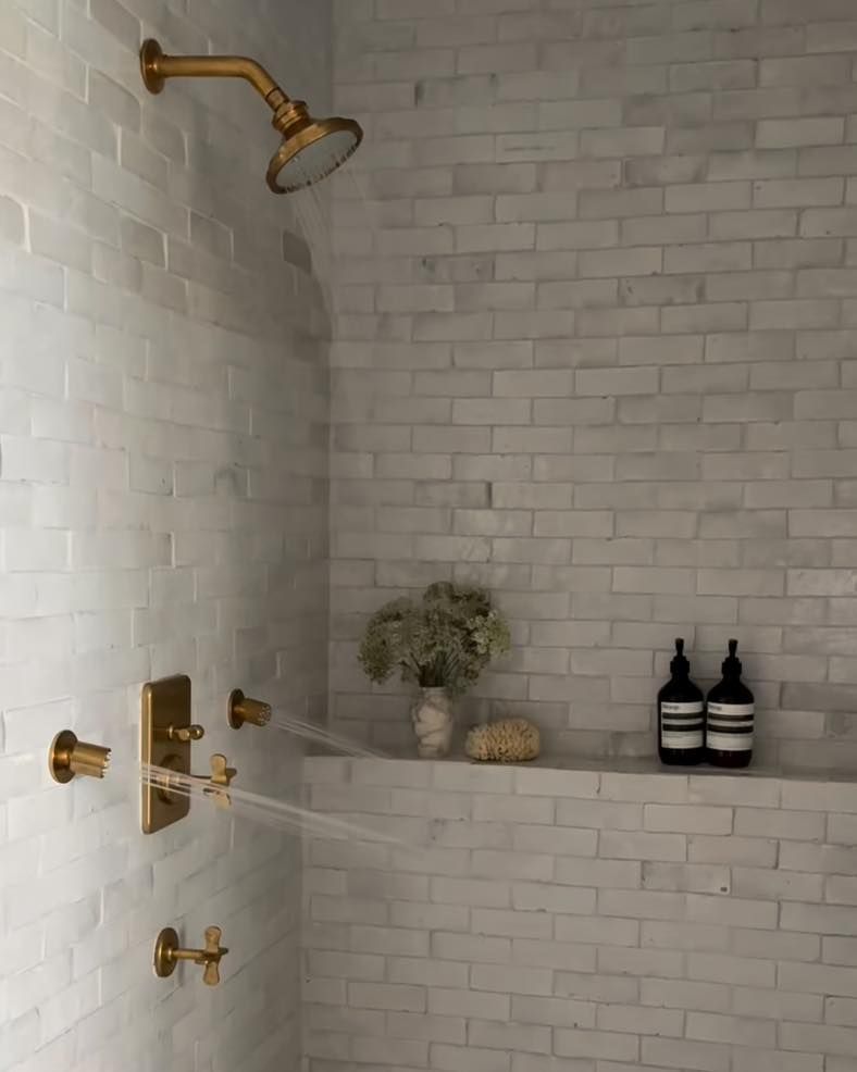 Gold shower fixtures and white brick-like tile in a minimalist bathroom; small shelf with toiletries.