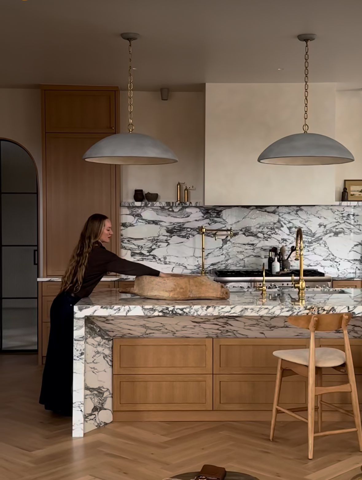 Woman leans over marble island in a modern kitchen with pendant lights and wood cabinetry.