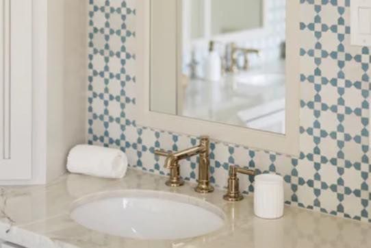 Bathroom sink with gold faucet, white countertop, and patterned blue and white tile backsplash.
