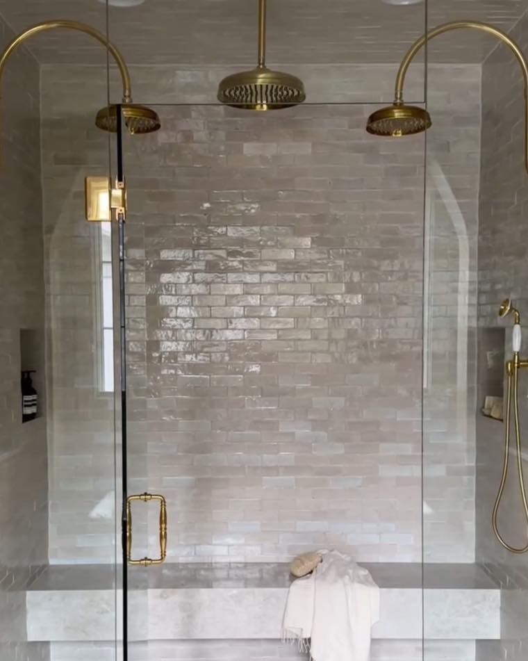 Shower with three gold showerheads against light brick tile, glass doors, and a built-in bench.