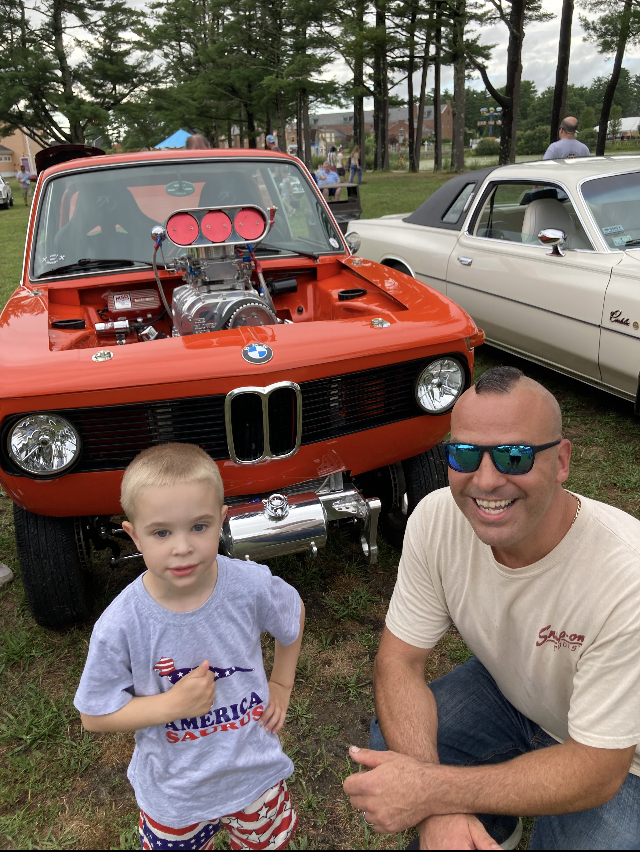 A man and a little boy are posing for a picture in front of an orange car.