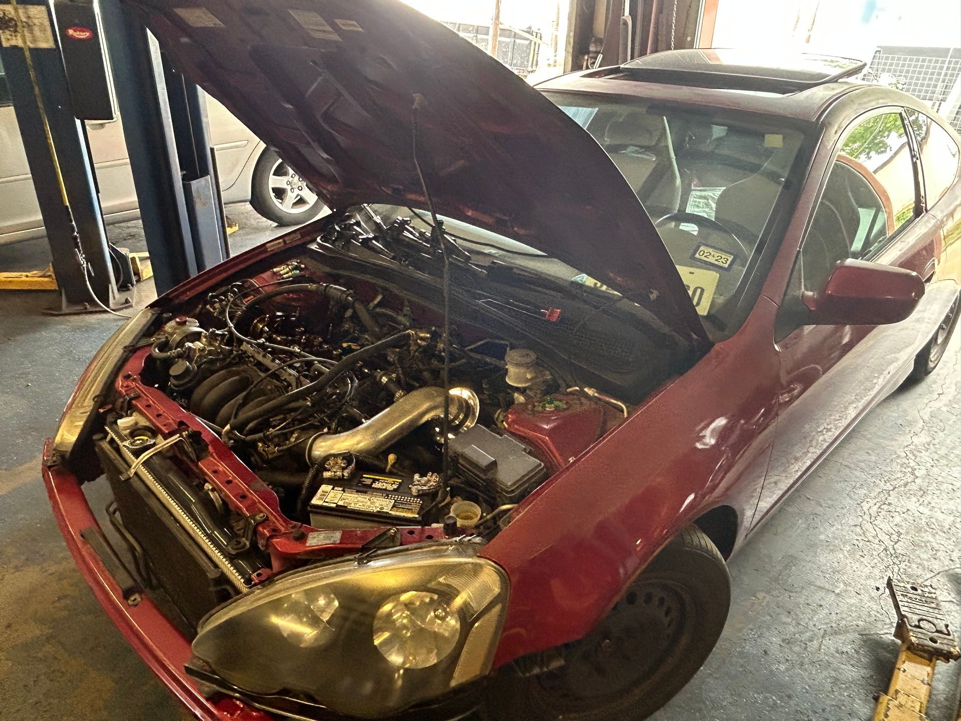 Red car with open hood in a garage, revealing engine components. Sunroof visible.