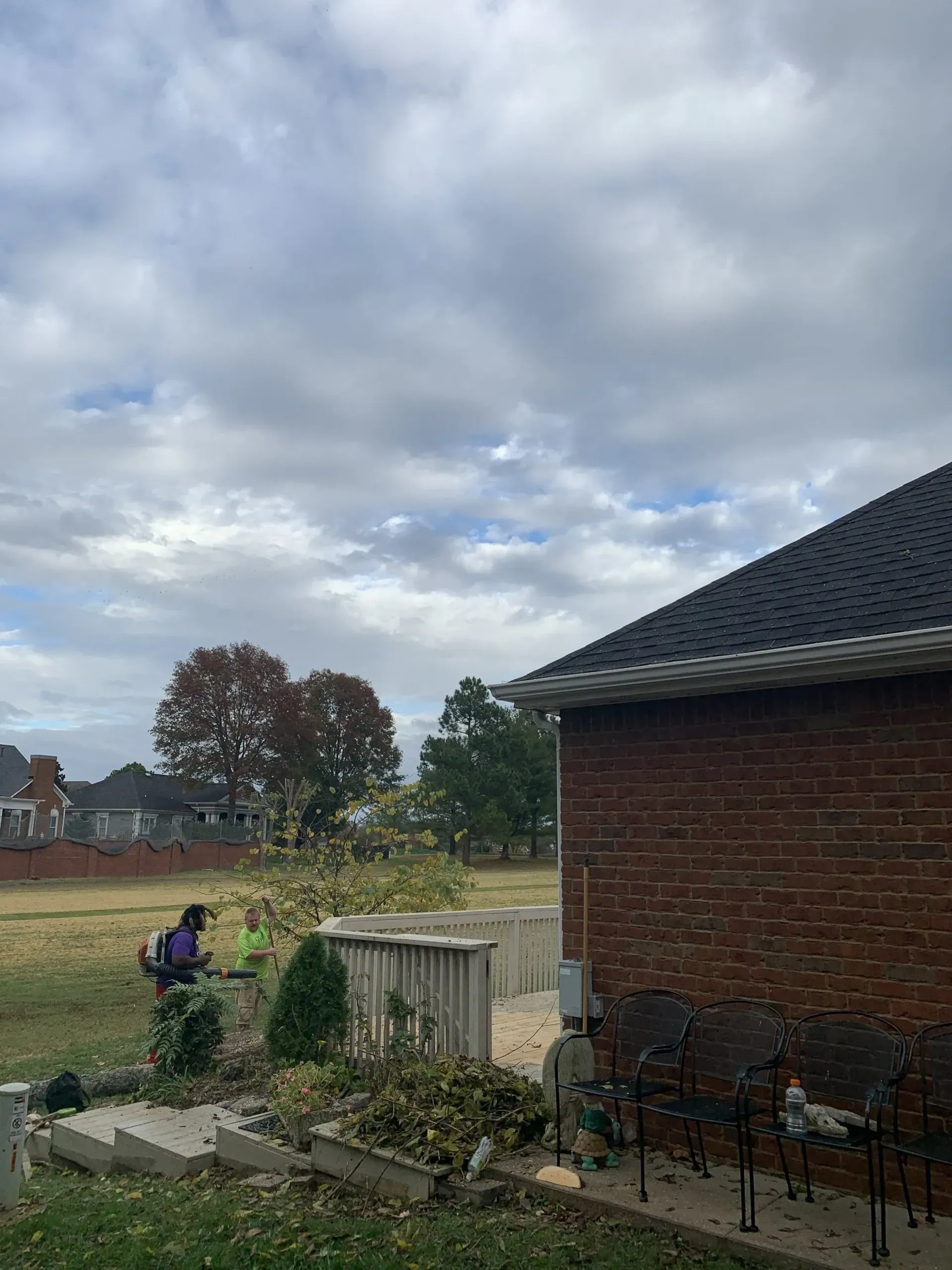 Person trimming bushes on a cloudy day; brick house on the right.