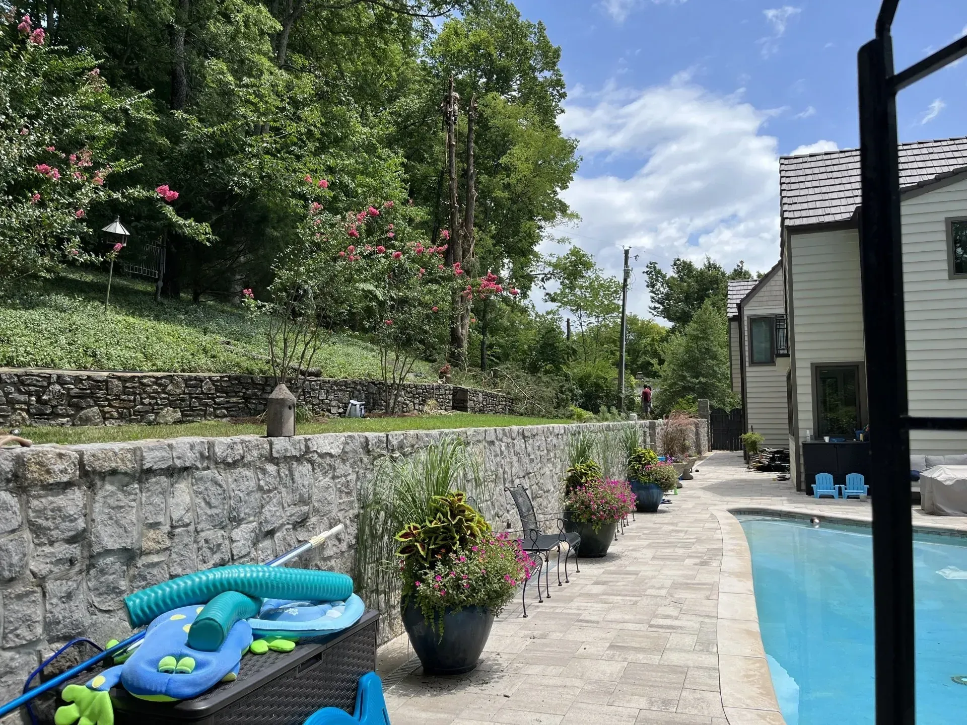 Backyard with a stone wall, pool, and lush greenery on a sunny day.