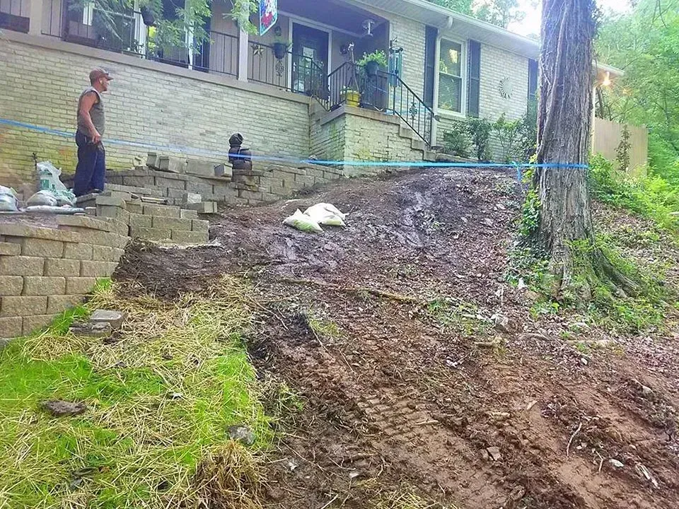 Workers build retaining wall and steps at a house entrance. Muddy hill beside tree; blue guide line.
