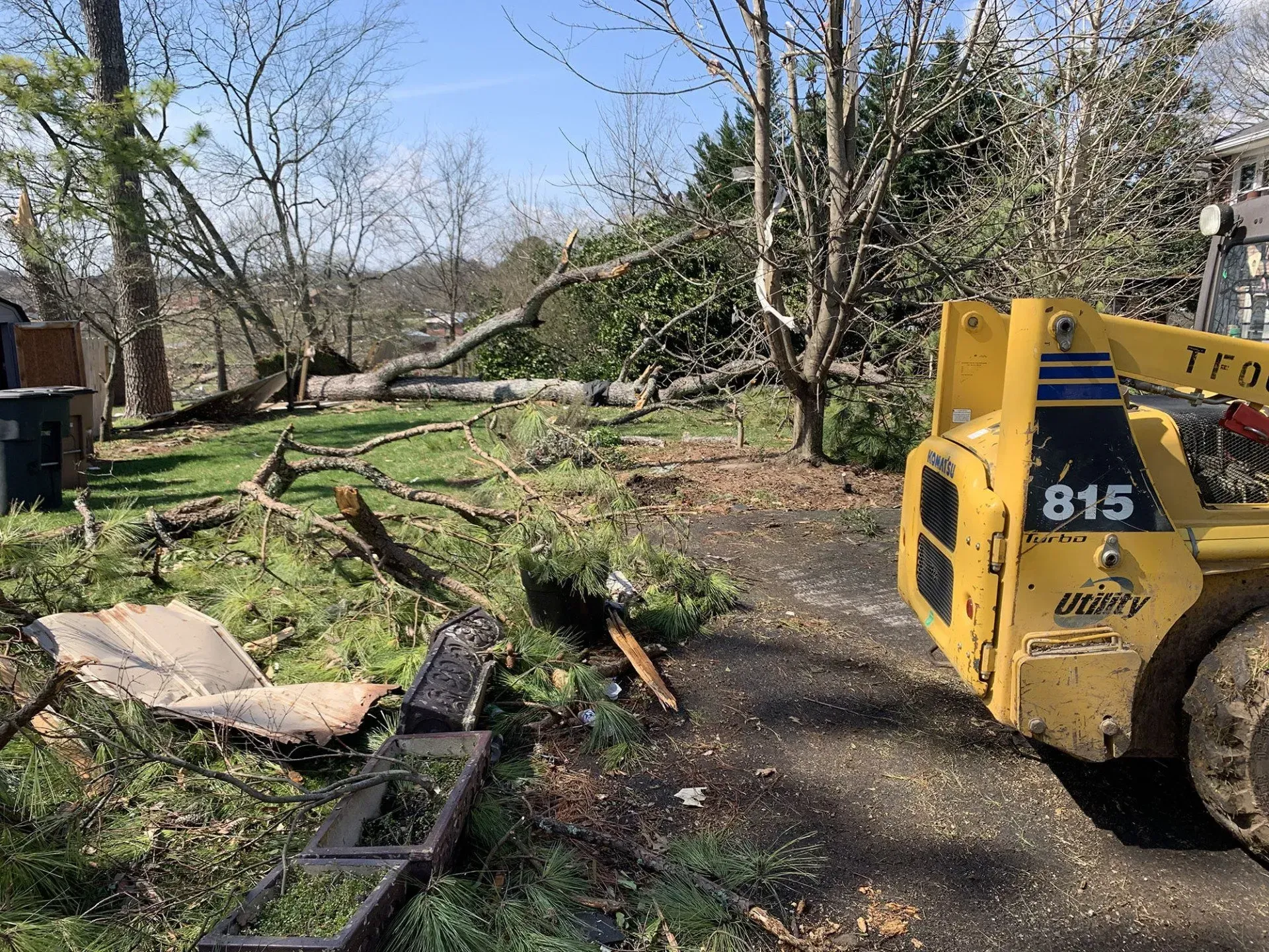 Yellow skid steer clears debris from a yard with fallen trees and a damaged structure.