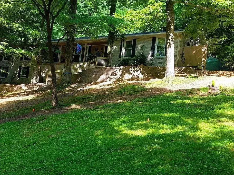 Ranch-style house nestled among trees with a sloped yard. Green grass and foliage surround the building.