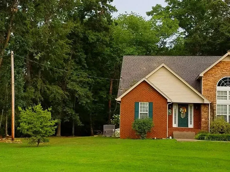 Brick house with green shutters and front door, set on a grassy lawn, backed by a forest.