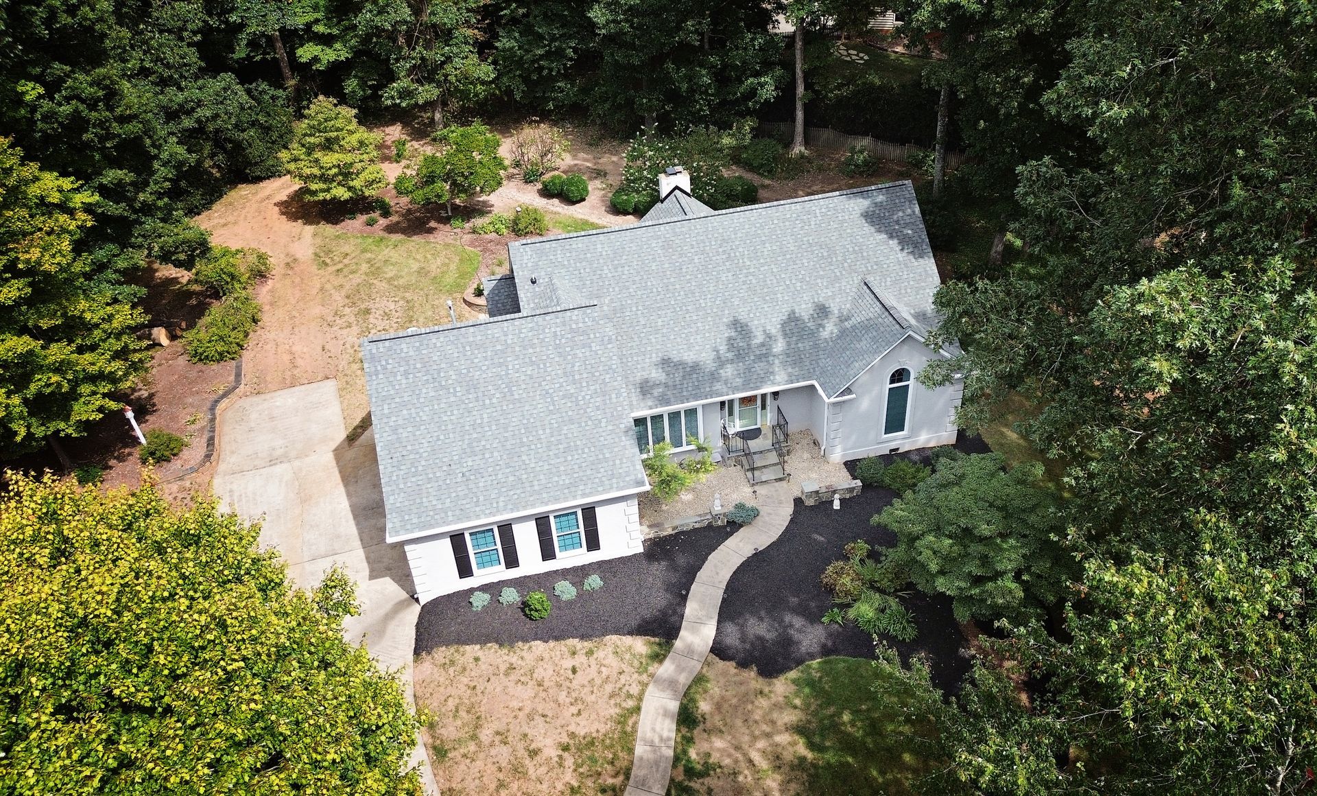 Aerial view of a gray house with a dark roof surrounded by trees and a paved driveway.