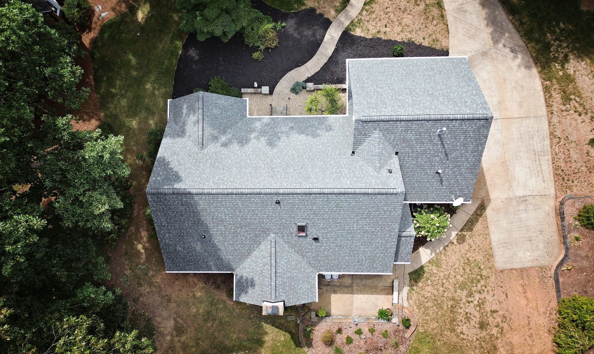 Aerial view of a gray-roofed house surrounded by trees, a driveway, and a small courtyard.
