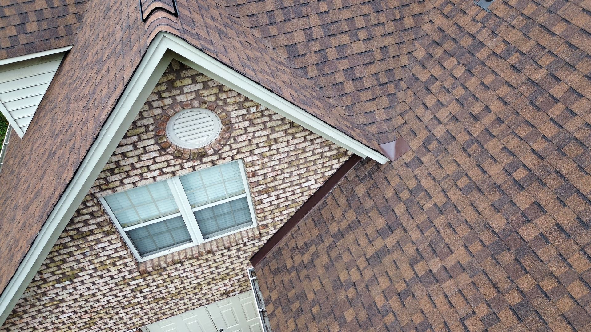 Brown shingle roof on a brick house with a white-framed window and a circular vent.