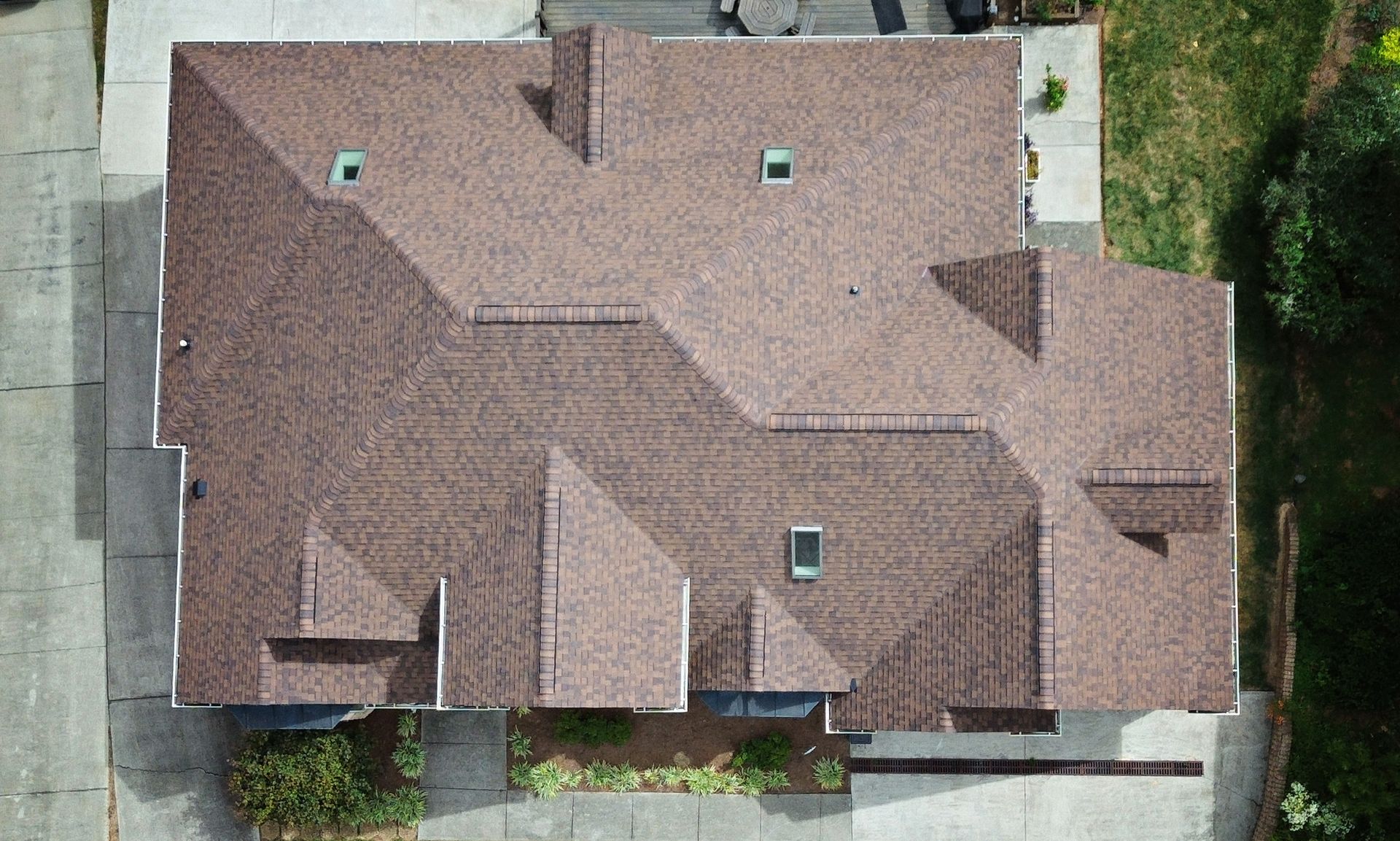 Overhead view of a multi-sectioned brown shingle roof on a house, set on concrete and next to greenery.