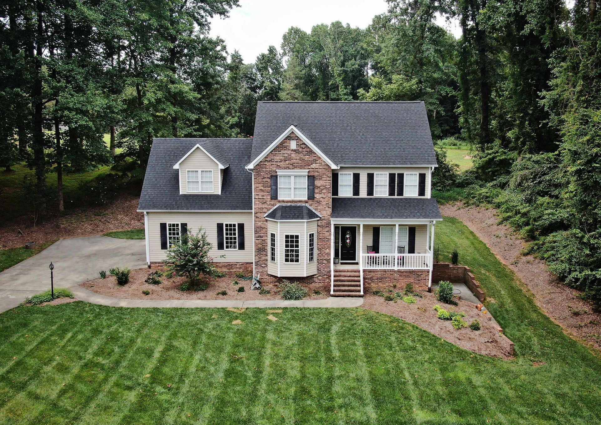Two-story house with brick facade, black shutters, white trim, and a manicured lawn surrounded by trees.