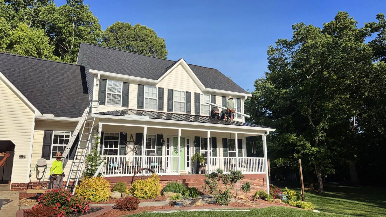 House with workers on the roof, ladder on the side. Blue sky, green trees, beige siding, white trim.
