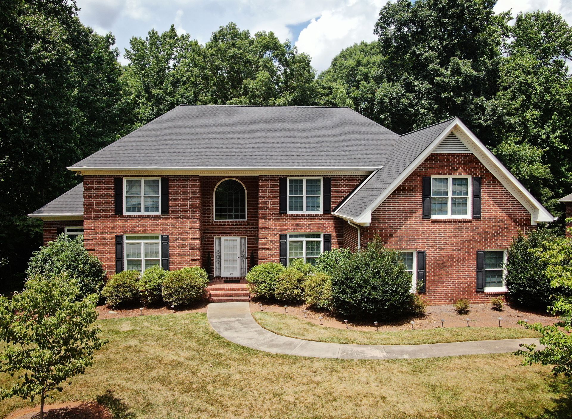 Brick two-story house with black shutters and a dark gray roof, surrounded by green trees and bushes.