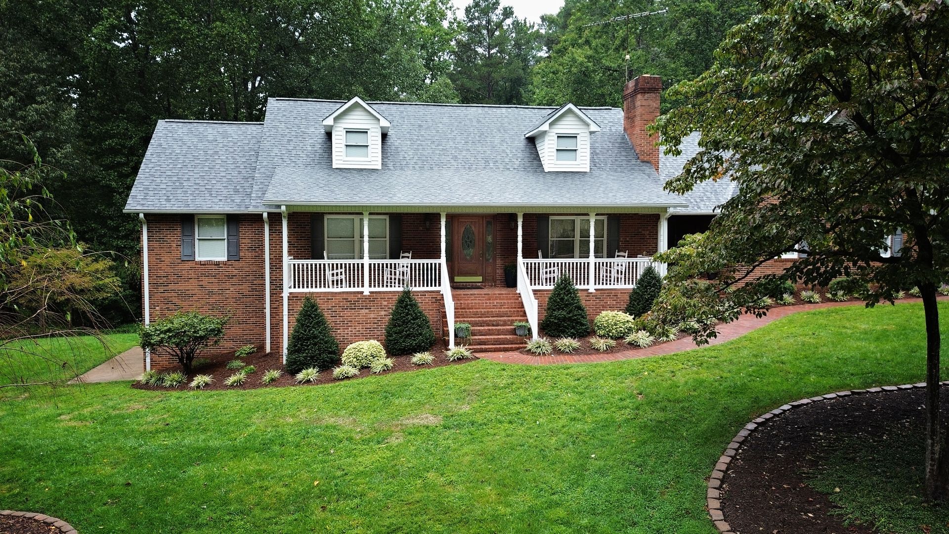 Brick house with porch, dormers, and green lawn. Landscaping with shrubs and trees.
