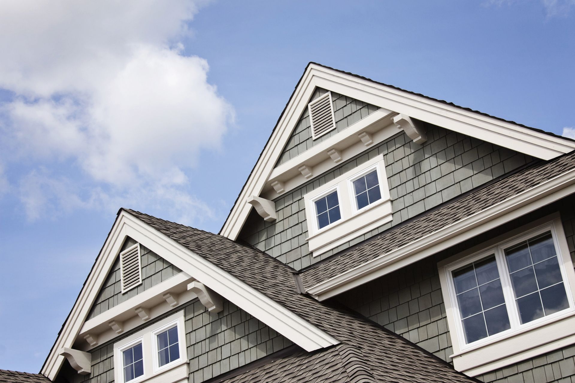 Green shingled house with white trim against a blue sky, featuring gables and windows.