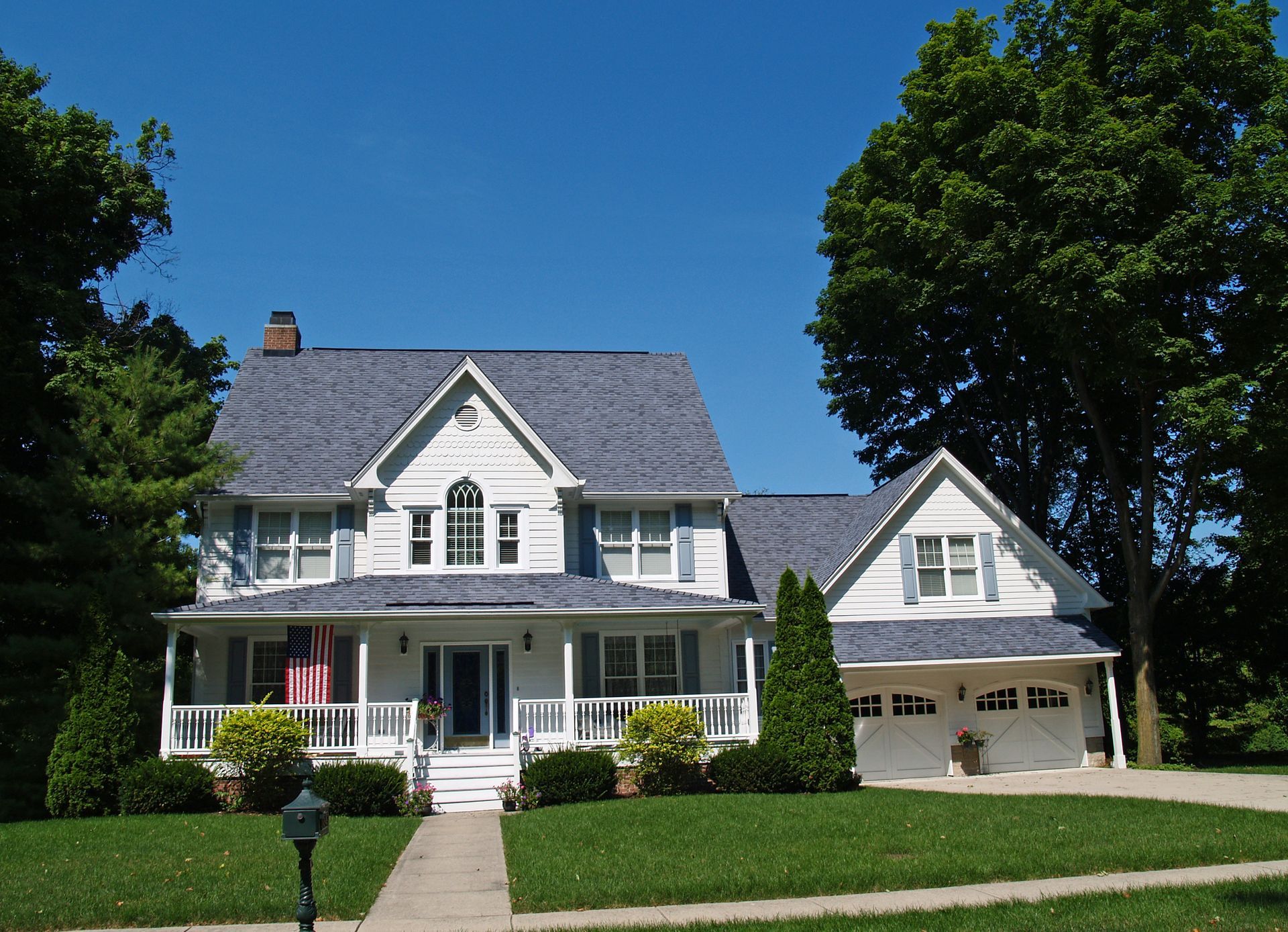 White two-story house with blue roof and shutters. Porch, lawn, and trees under a clear, blue sky.