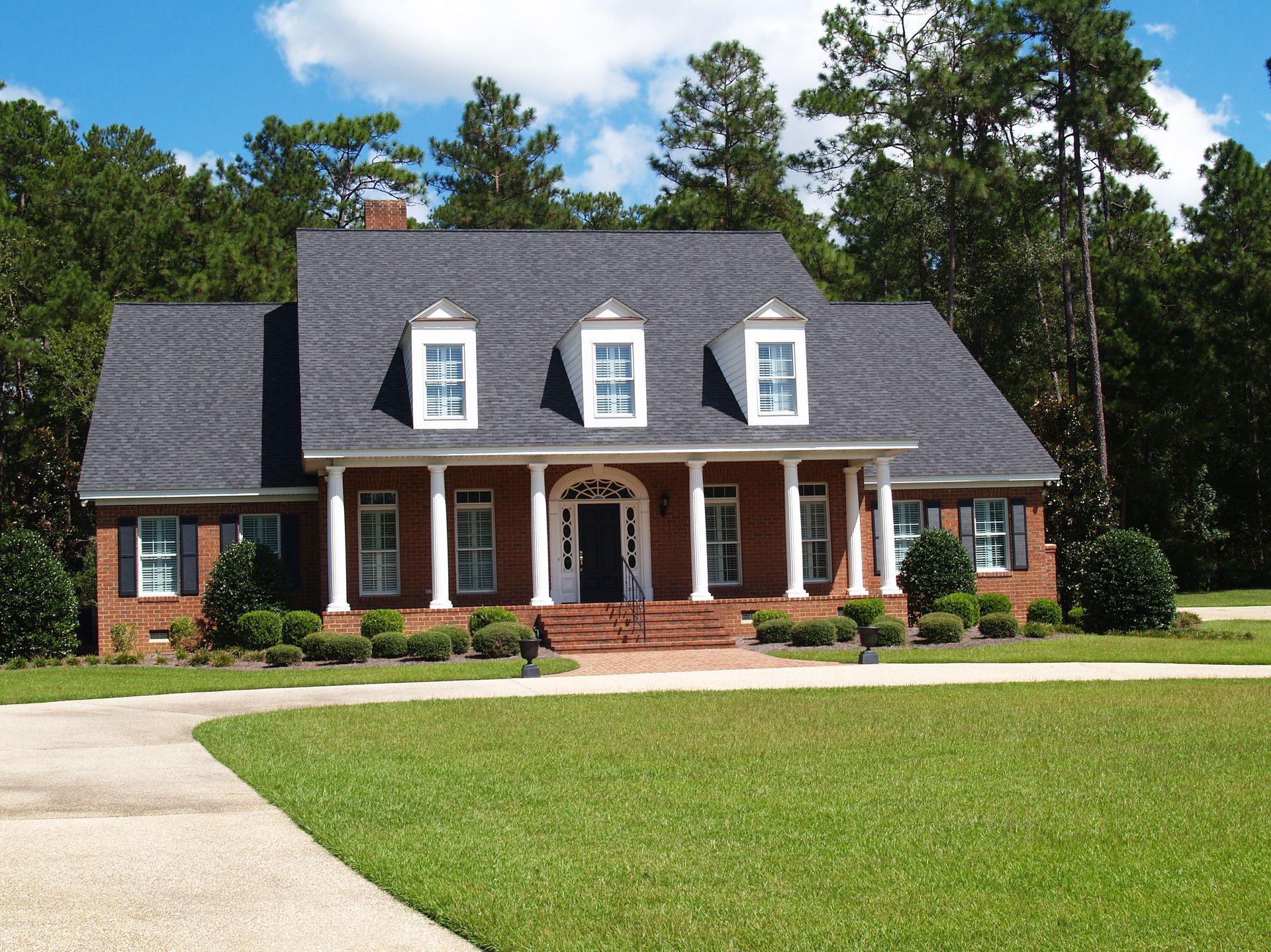 Brick house with white columns, dormers, and a dark shingle roof. Green lawn and driveway.