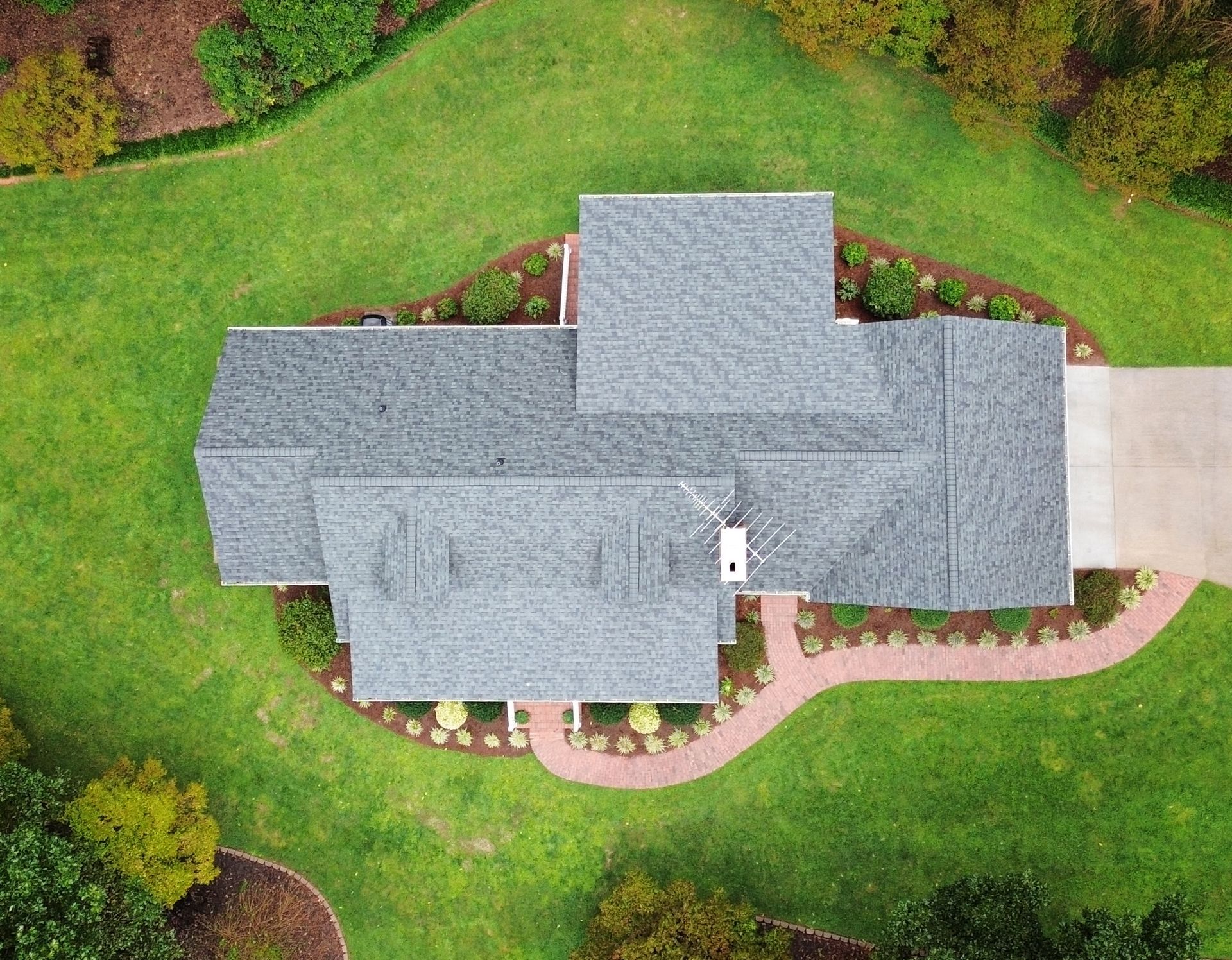 Overhead view of a gray-roofed house surrounded by a green lawn and landscaping. A concrete driveway is present.