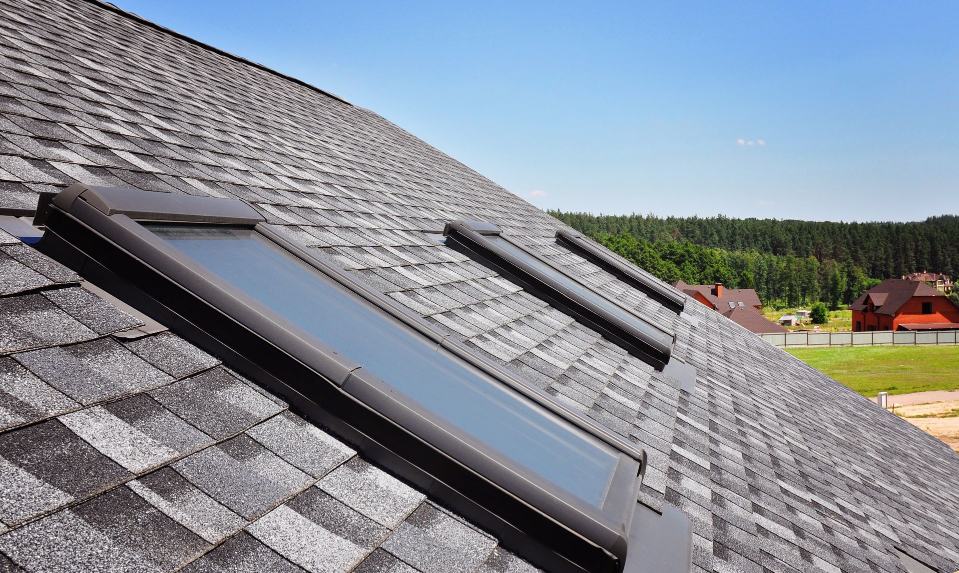 Gray shingle roof with three skylights, against a blue sky with trees and houses in the background.