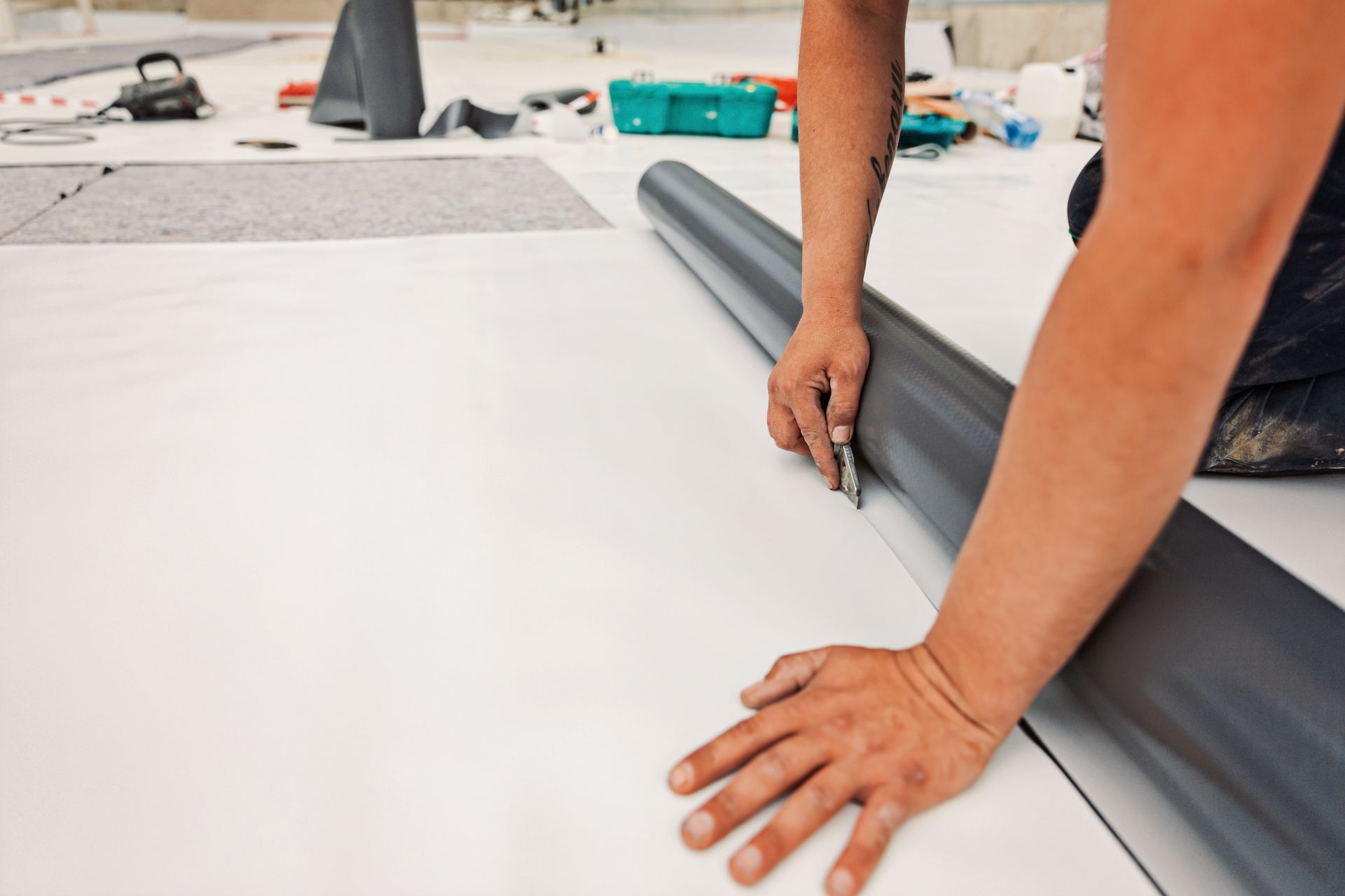 Person cutting a roll of dark flooring material on a white surface, with tools in the background.