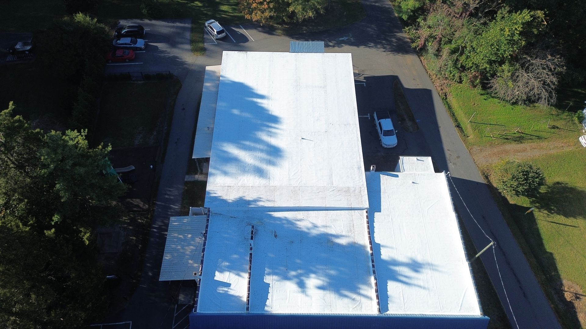 Aerial view of a white-roofed building. Cars are parked nearby, and green trees surround the building.