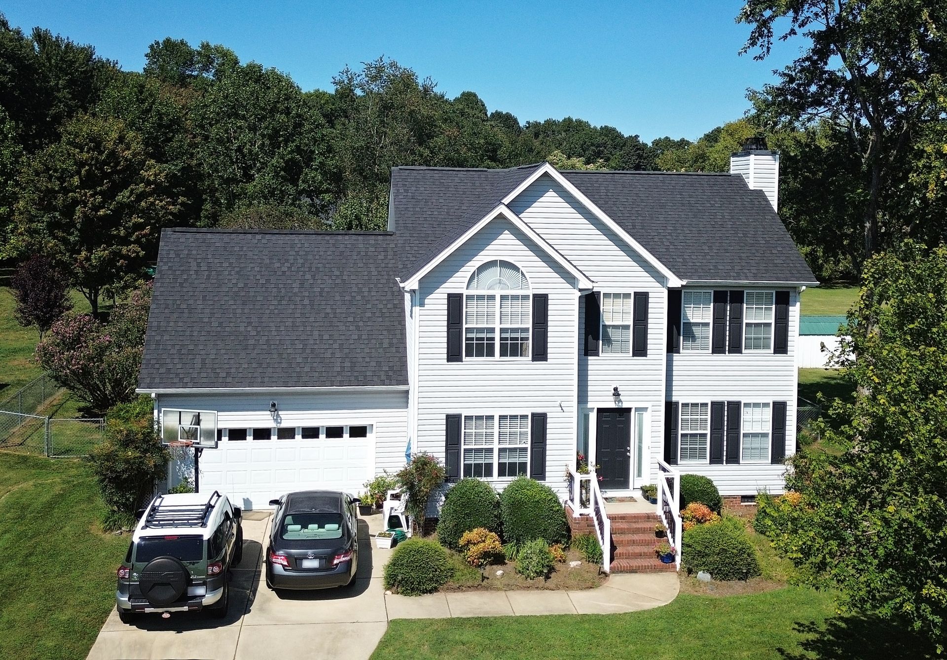 Two-story white house with black shutters, a dark roof, and two cars parked in the driveway.