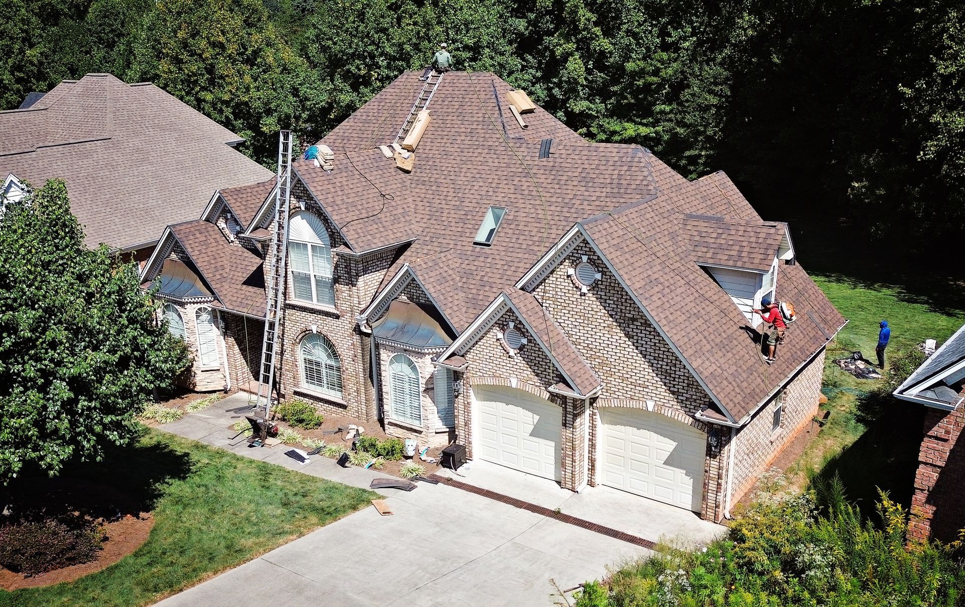 House with roofers working on a brown shingle roof; green lawn and trees.