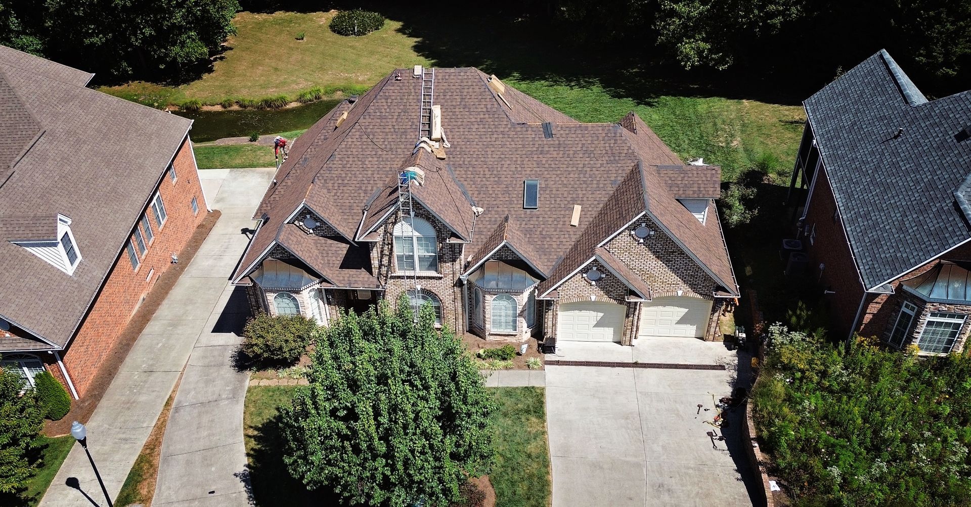 Aerial view of a large house with brown roof, stone facade, and two-car garage. Houses flank it.