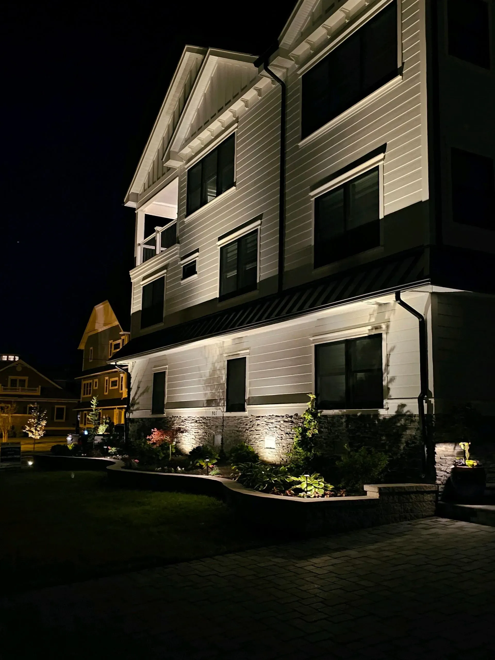 Two-story white house at night, lit by spotlights. Garden with plants, another house visible in the distance.