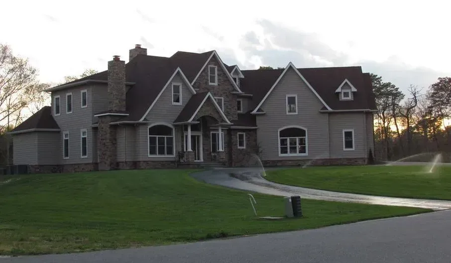 Large two-story house with gray siding, stone accents, and brown roof; on a green lawn with a driveway.