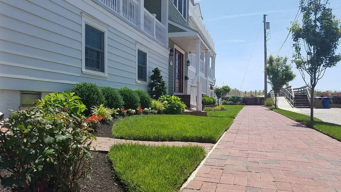 A white house with a porch and red door, next to a brick walkway and green lawn under a sunny sky.