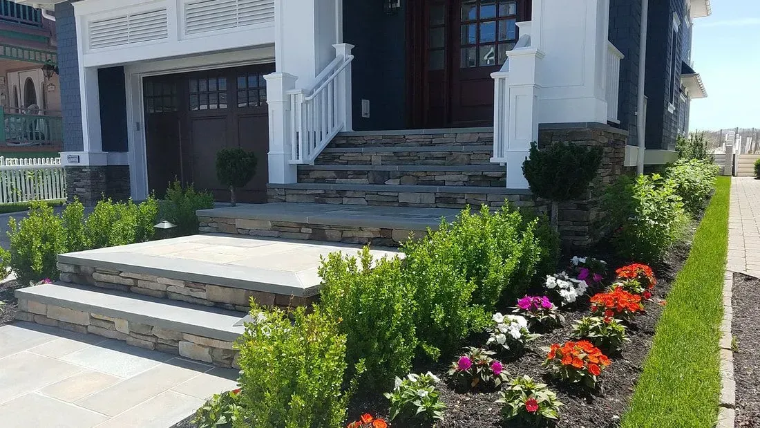 Stone steps lead to a blue house with white trim. Landscaping includes green bushes and colorful flowers.