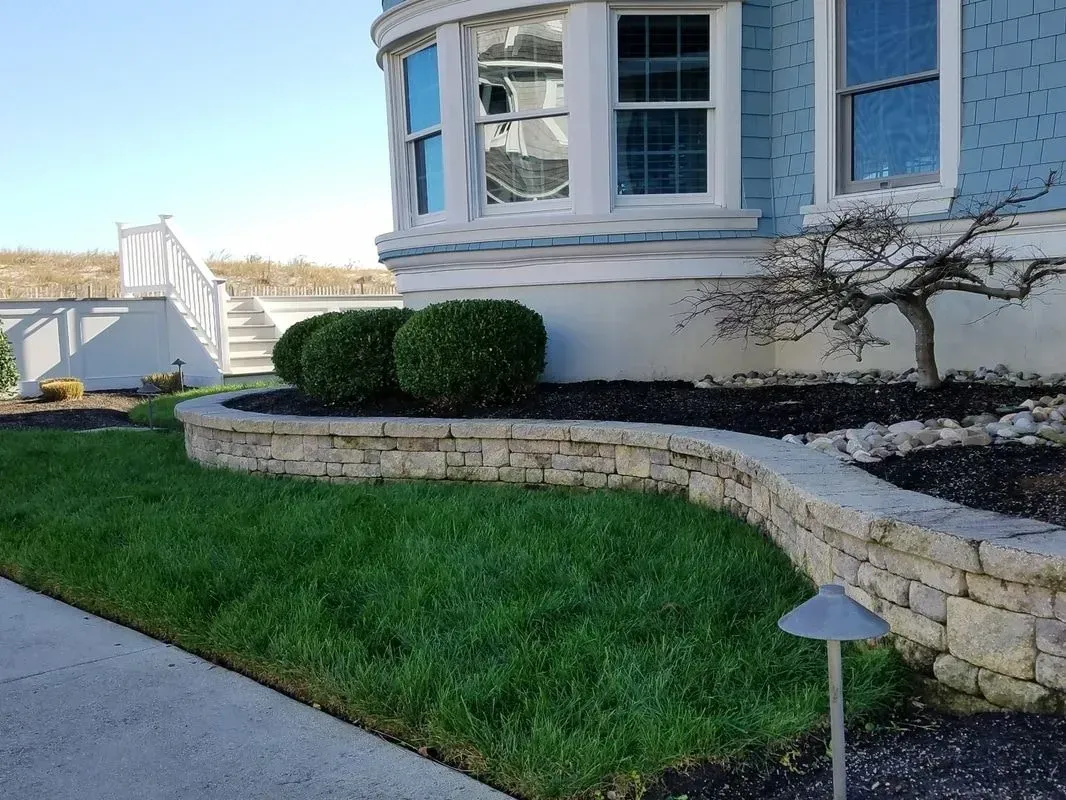 Stone retaining wall and green lawn in front of a blue house with bay windows.