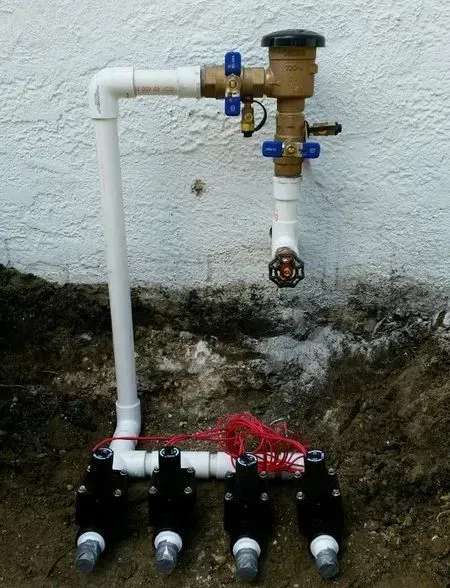 Irrigation system with PVC pipes, brass valve, and four black solenoid valves against a white wall and dirt.